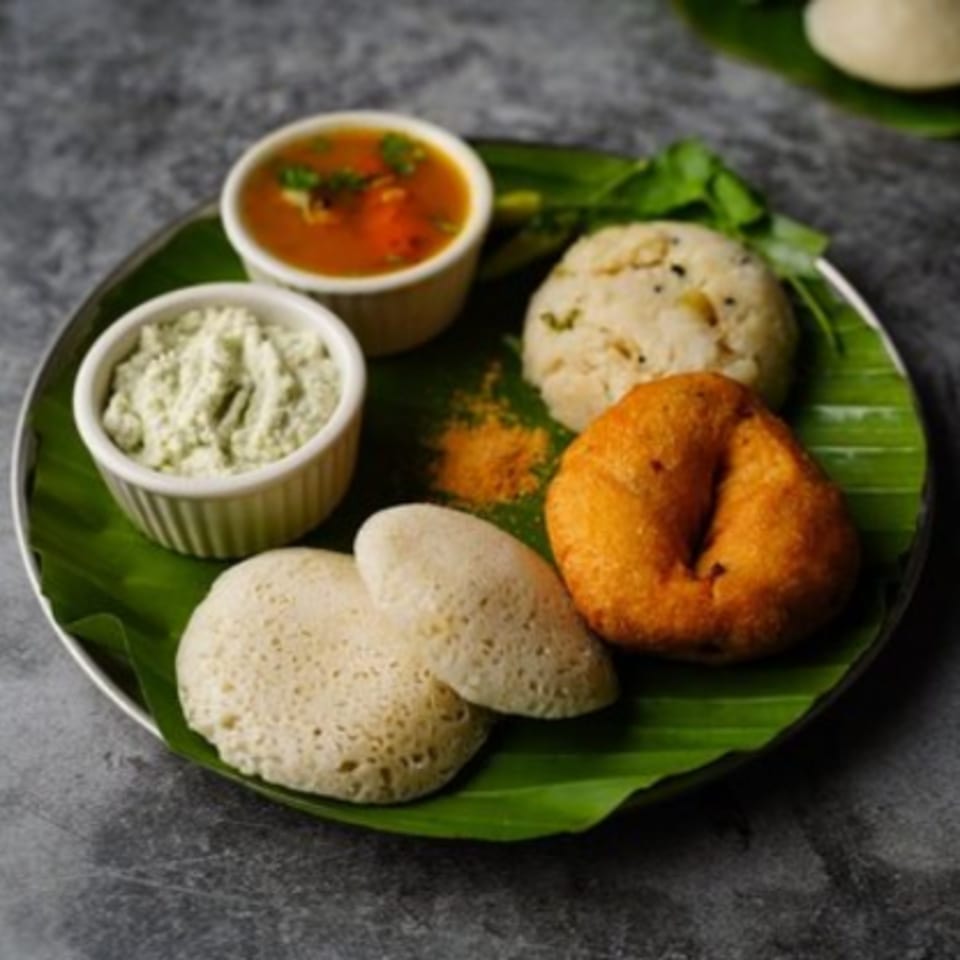 A plate of Indian food with idli, vada, upma, sambar, and chutney on a banana leaf.