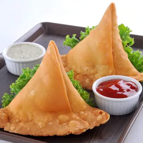 Two golden-brown samosas with lettuce, served with mint chutney and ketchup on a dark tray.