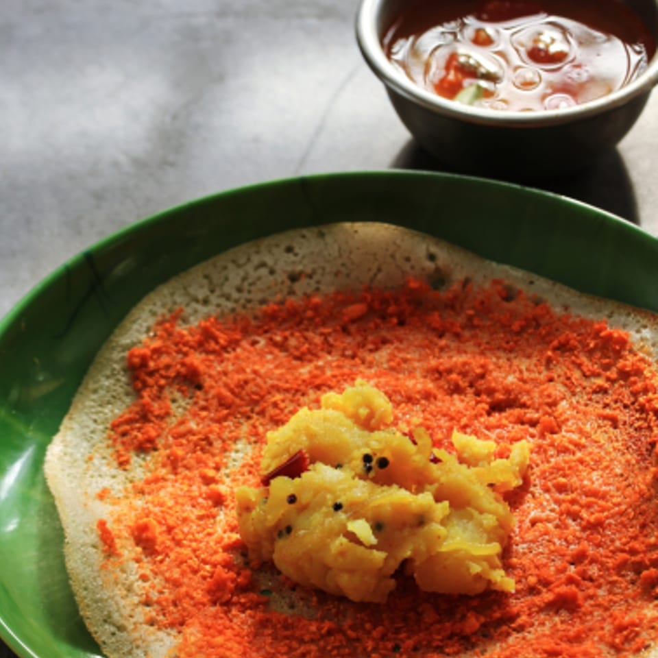 A dosa on a green plate, topped with orange powder and a scoop of yellow potato masala, with sambar.