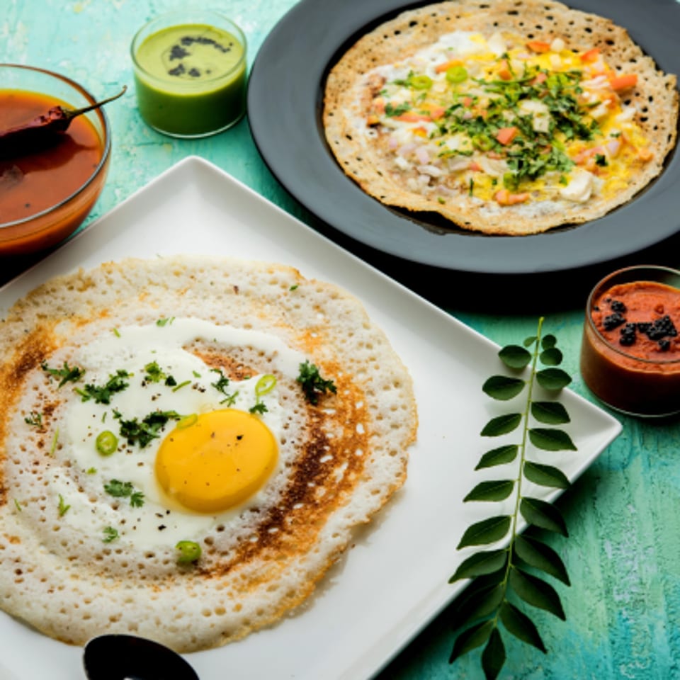 Two Indian dosas with egg, one with a fried egg, served with chutneys and sambar.